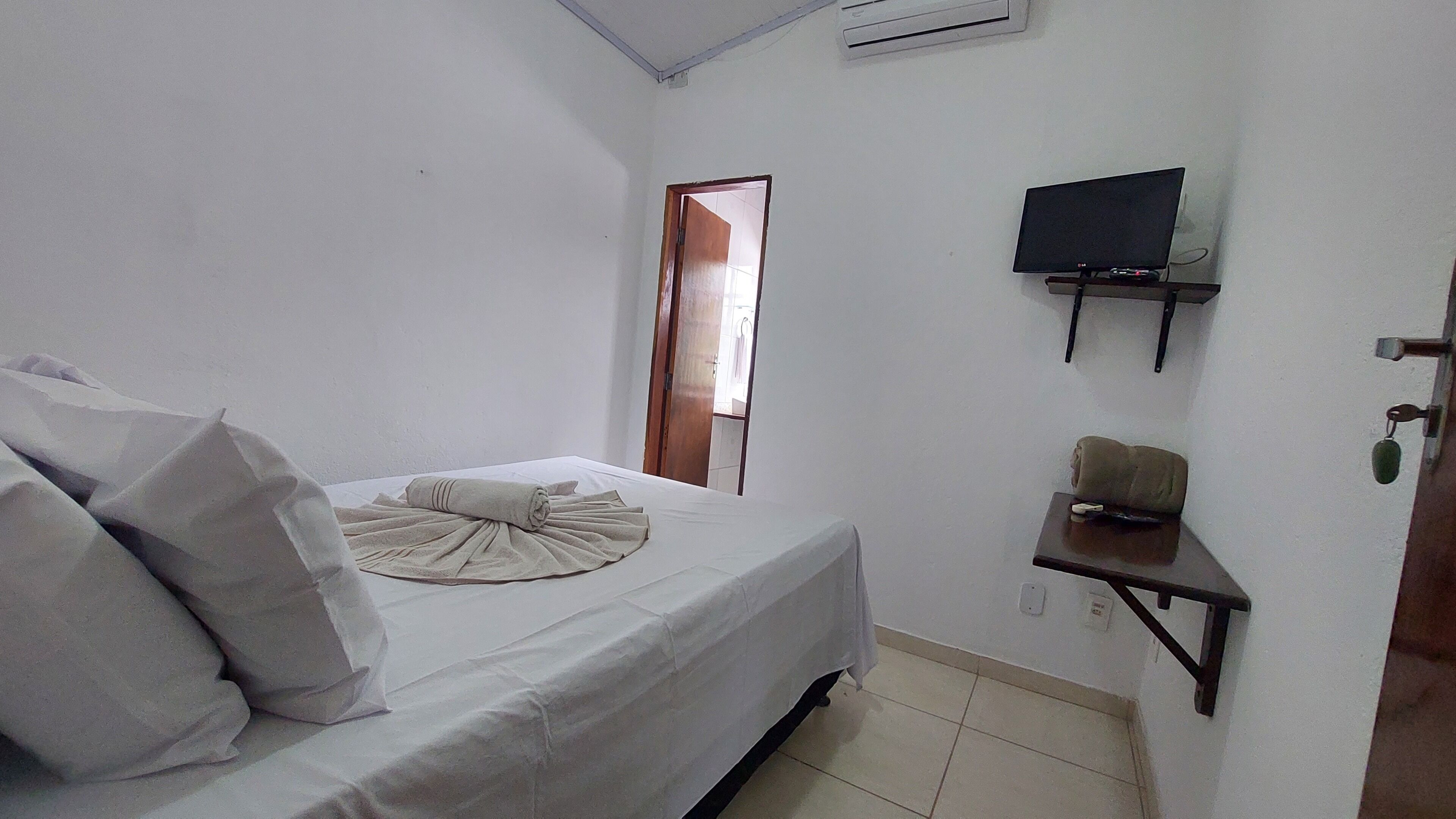 A bedroom with a bed, pillows, a TV on a stand, a door, and a wall-mounted shelf.