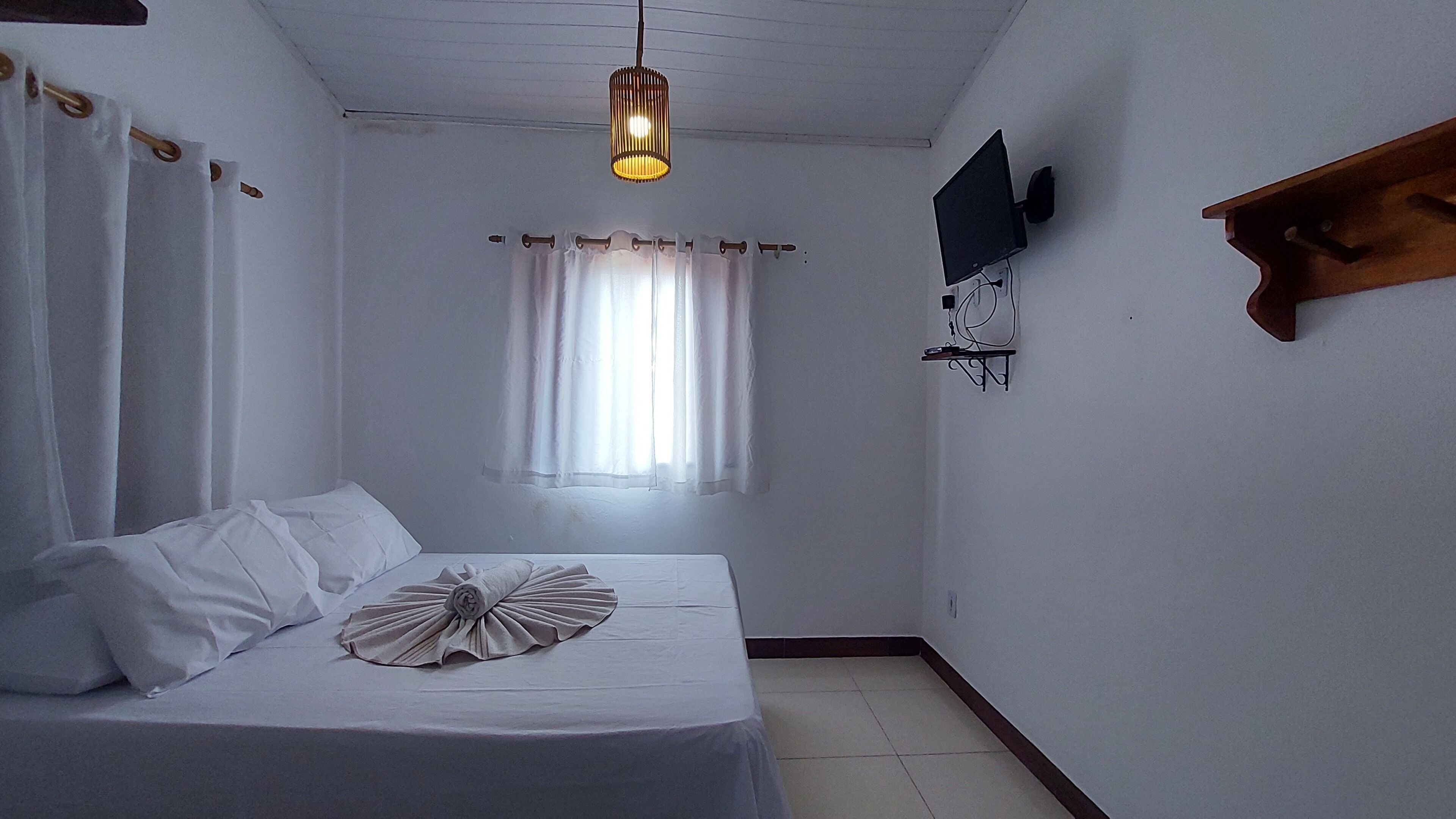 A bedroom with a bed, curtains, a hanging light fixture, a mounted TV, and a wooden shelf.