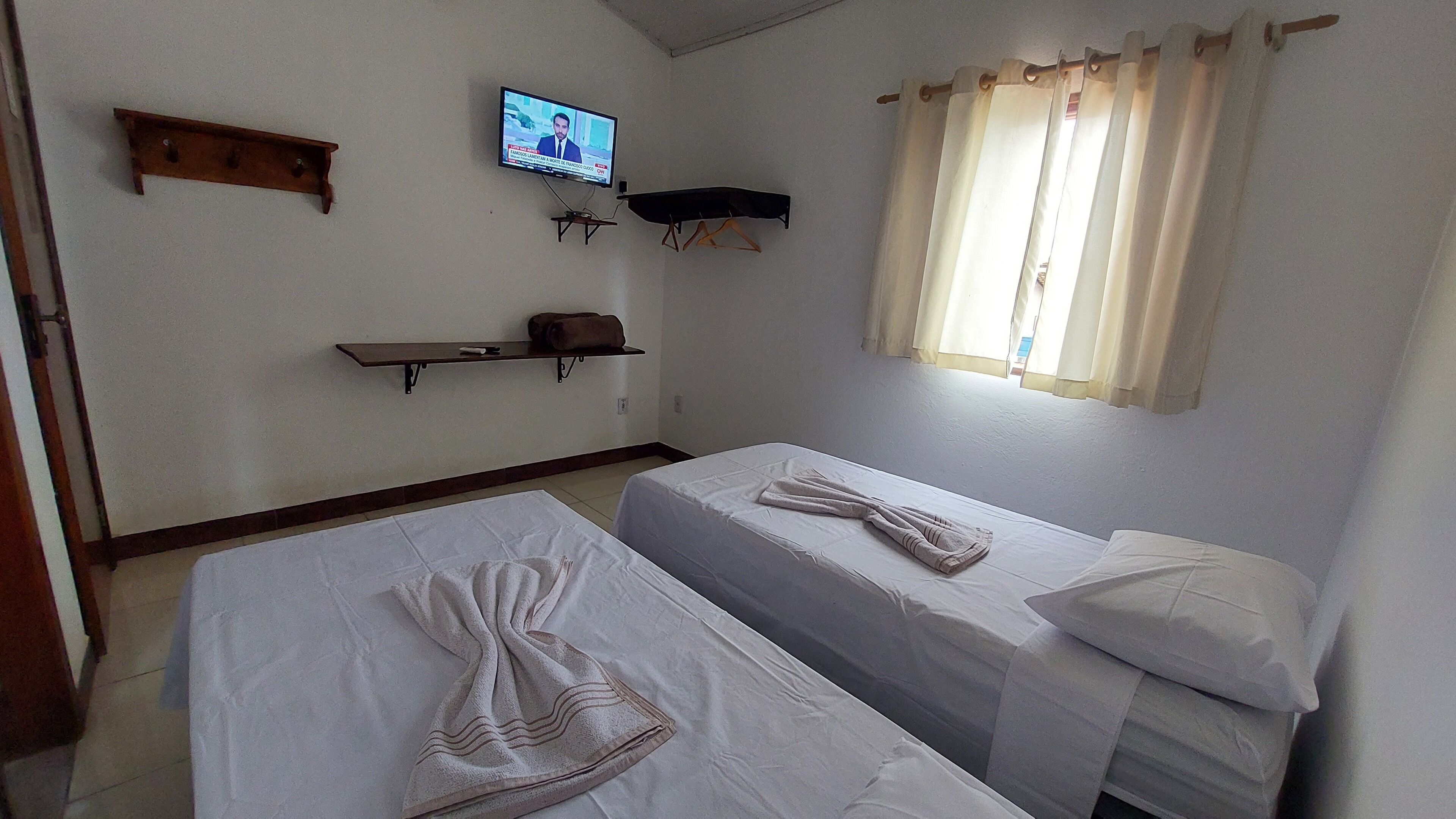 A room with two beds, a TV on the wall, a window with curtains, and a shelf with a bag.