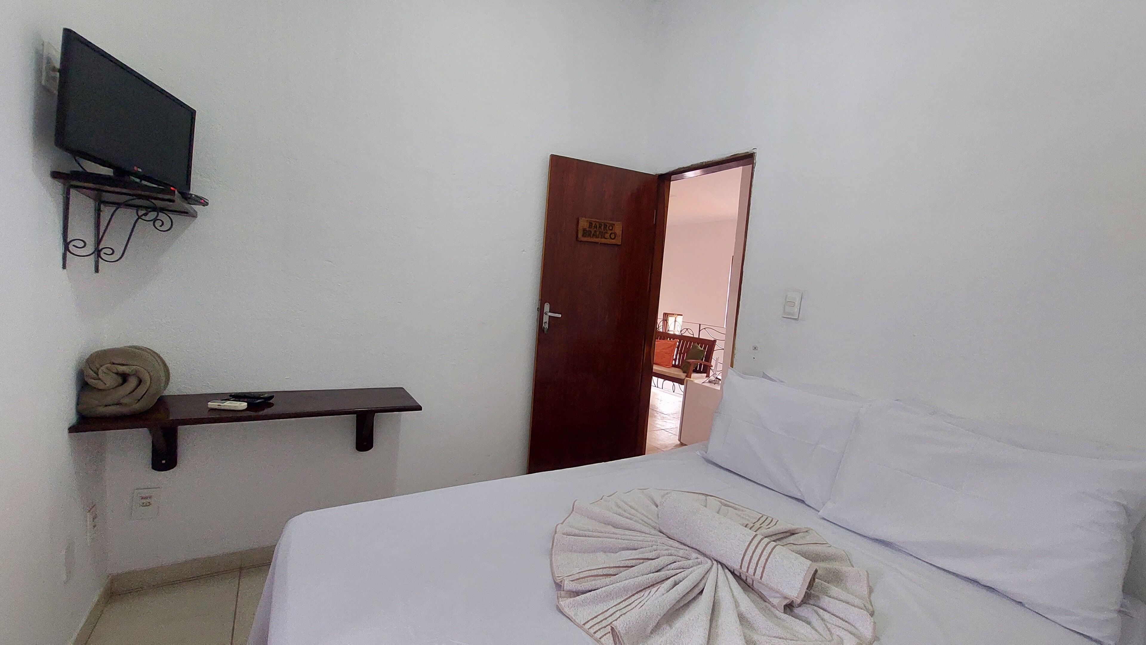 Un dormitorio con cama, televisor en soporte de pared, puerta de madera y una pequeña estantería con una toalla y un control remoto.