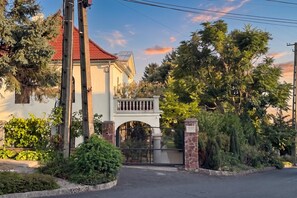 A gated entrance to a building with a red roof, surrounded by lush greenery and trees. - Balchill House With Sauna And Jacuzzi (Balatonszepezd)