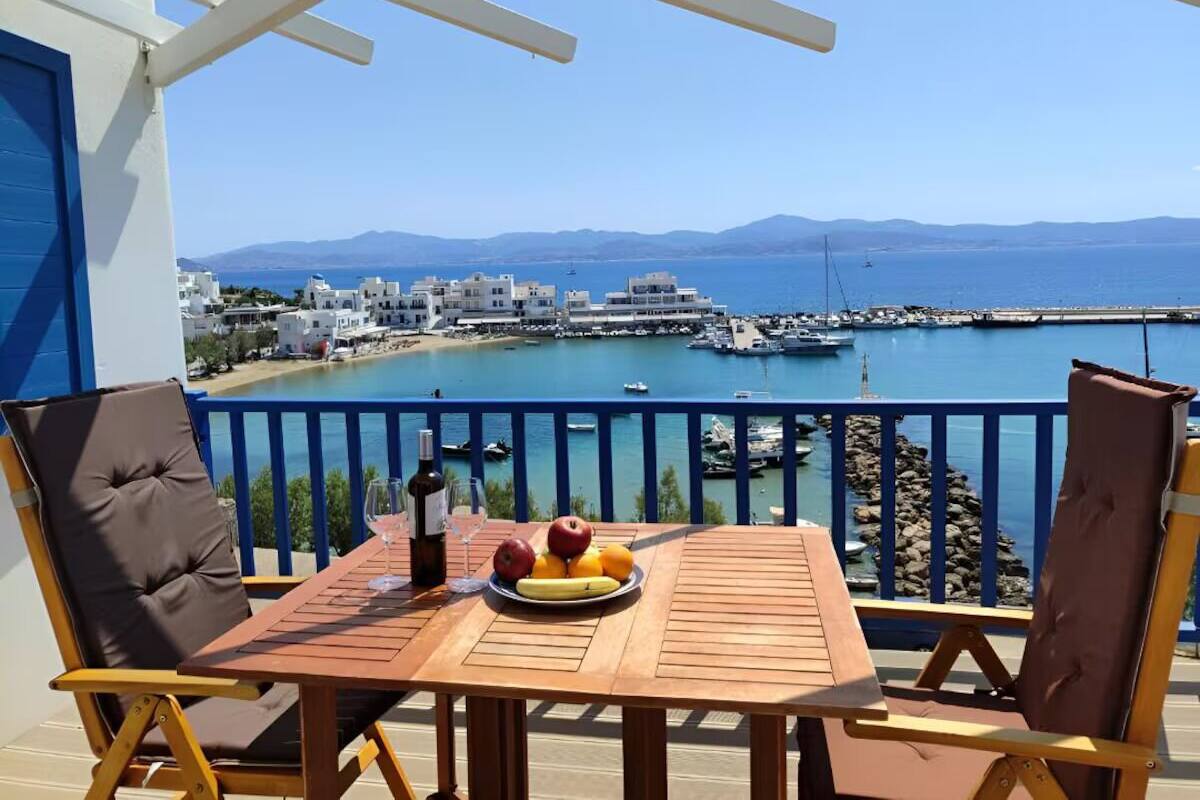 Une table en bois, sur laquelle sont disposés un plateau de fruits, une bouteille de vin et des verres, se trouve sur un balcon donnant sur une marina et la mer.