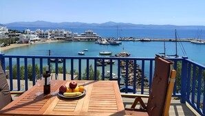 A wooden table with a fruit plate, a bottle, and glasses on a balcony overlooking a marina with boats and buildings in the background. - Hotel Corali (Paros)
