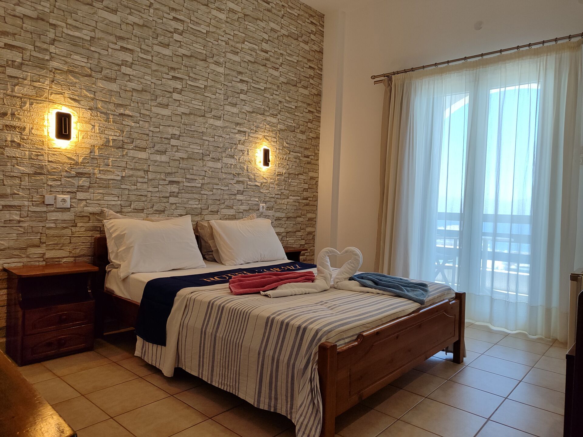 A hotel room with a stone wall, a bed with striped bedding, a wooden nightstand, and a view of the outdoors through sheer curtains.