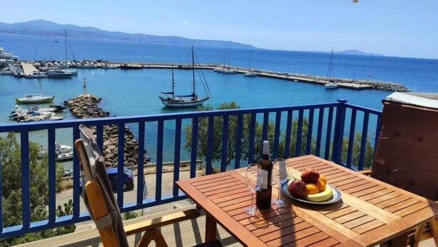 A wooden table with a bottle of wine, a glass, and fruit on a balcony overlooking a marina with boats and a clear blue sea. - Hotel Corali (Paros)