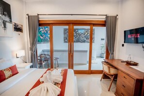 A hotel room with a bed, a desk, a chair, a TV, and a balcony with a painting. - Pondok Yogi Guest House Canggu (Canggu)