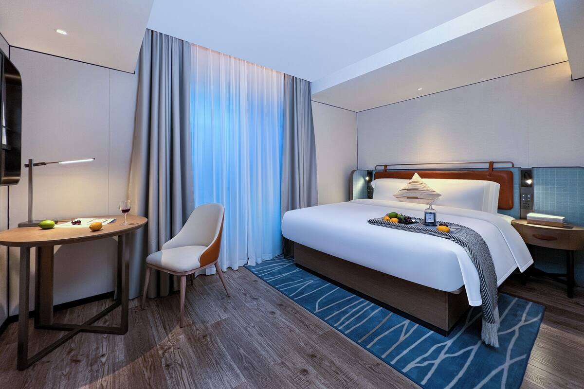 A modern hotel room with a large bed, a desk with a chair, a TV, and a window with curtains.
