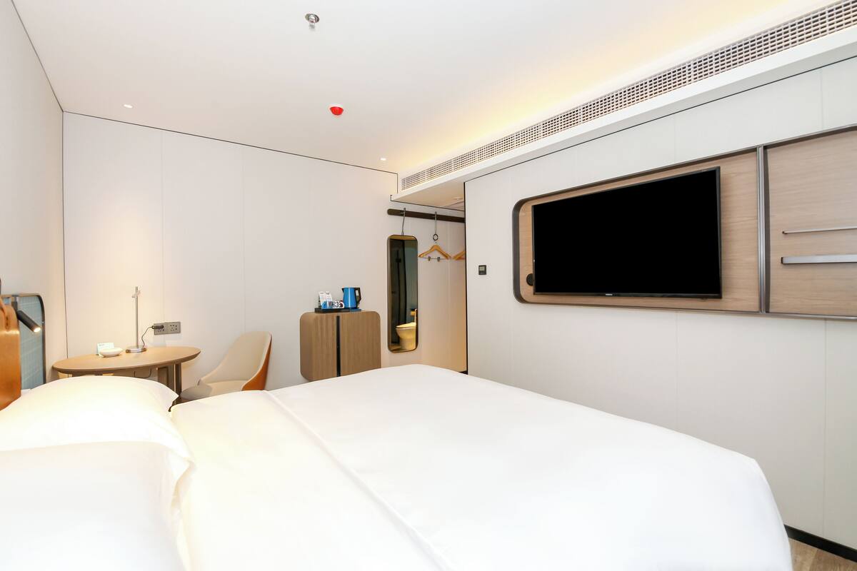 A hotel room with a large bed, a TV mounted on the wall, a small table with a lamp, and a wardrobe.