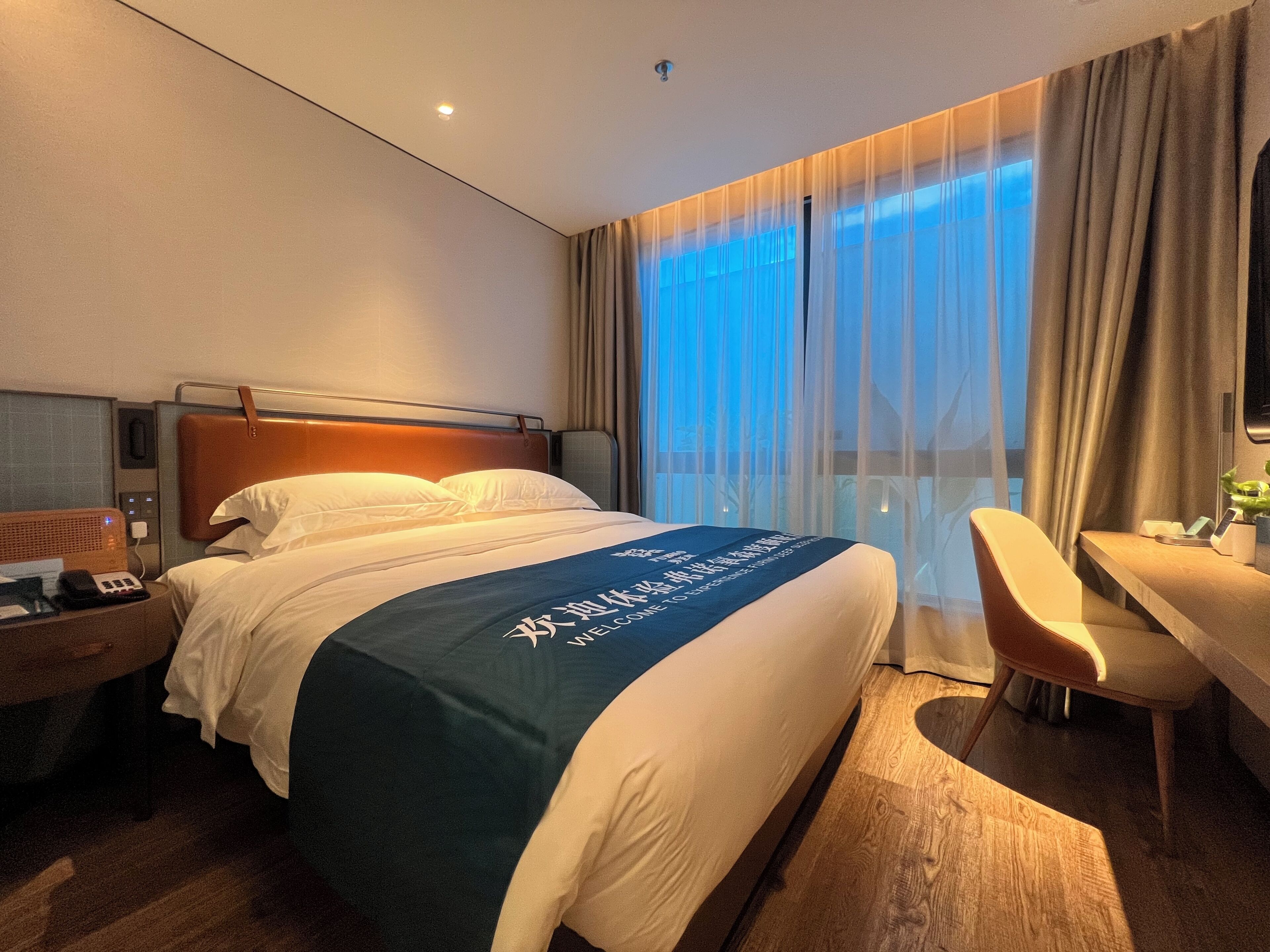 A hotel room with a bed, bedside table, chair, and a view of the outside through sheer curtains.