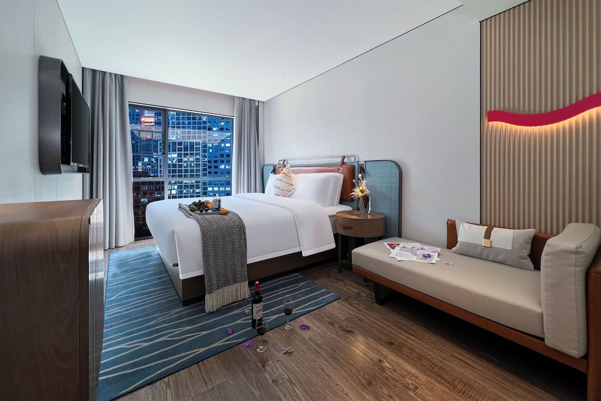 A modern hotel room with a large bed, a sofa, a TV, and a city view.