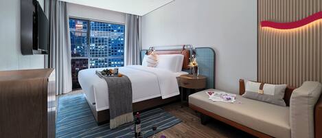 A modern hotel room with a large bed, a sofa, a TV, and a city view.