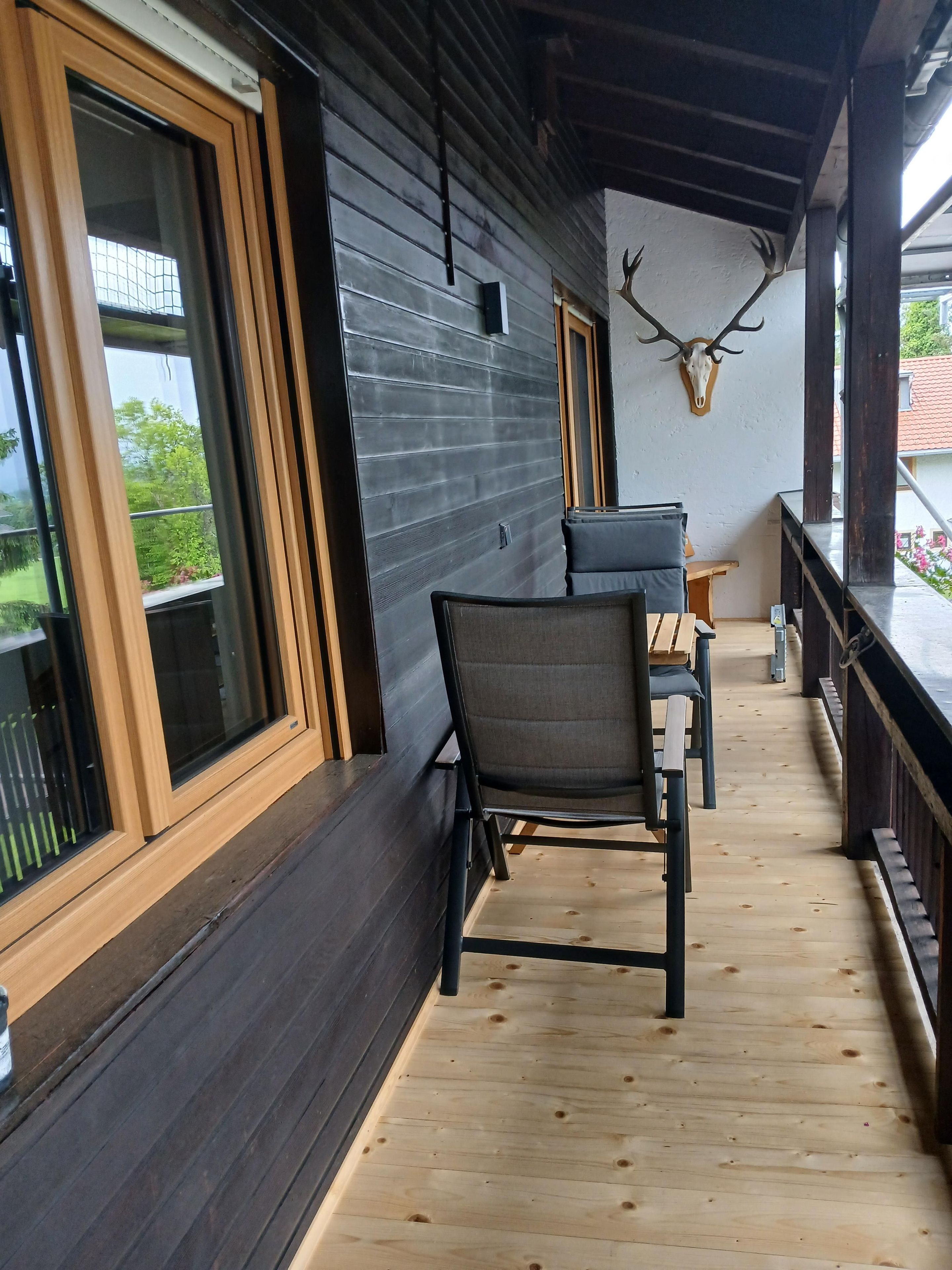 A wooden deck with a table and chairs, a mounted deer head, and a window overlooking greenery.