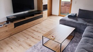 A modern living room with a flat-screen TV, wooden cabinets, a grey sofa, and a sliding glass door leading to an outdoor area. - Apartment 55 sqm for 2 persons with balcony and mountain view (Anger)