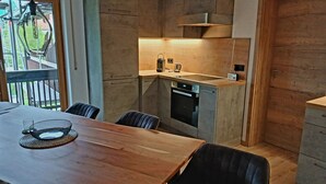 A modern kitchen with a built-in oven and microwave, a wooden dining table with chairs, and a view of the outdoors through a large window. - Apartment 55 sqm for 2 persons with balcony and mountain view (Anger)