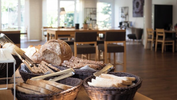 A bakery display with various breads and pastries in baskets, a wooden table, and a modern interior with chairs and tables.