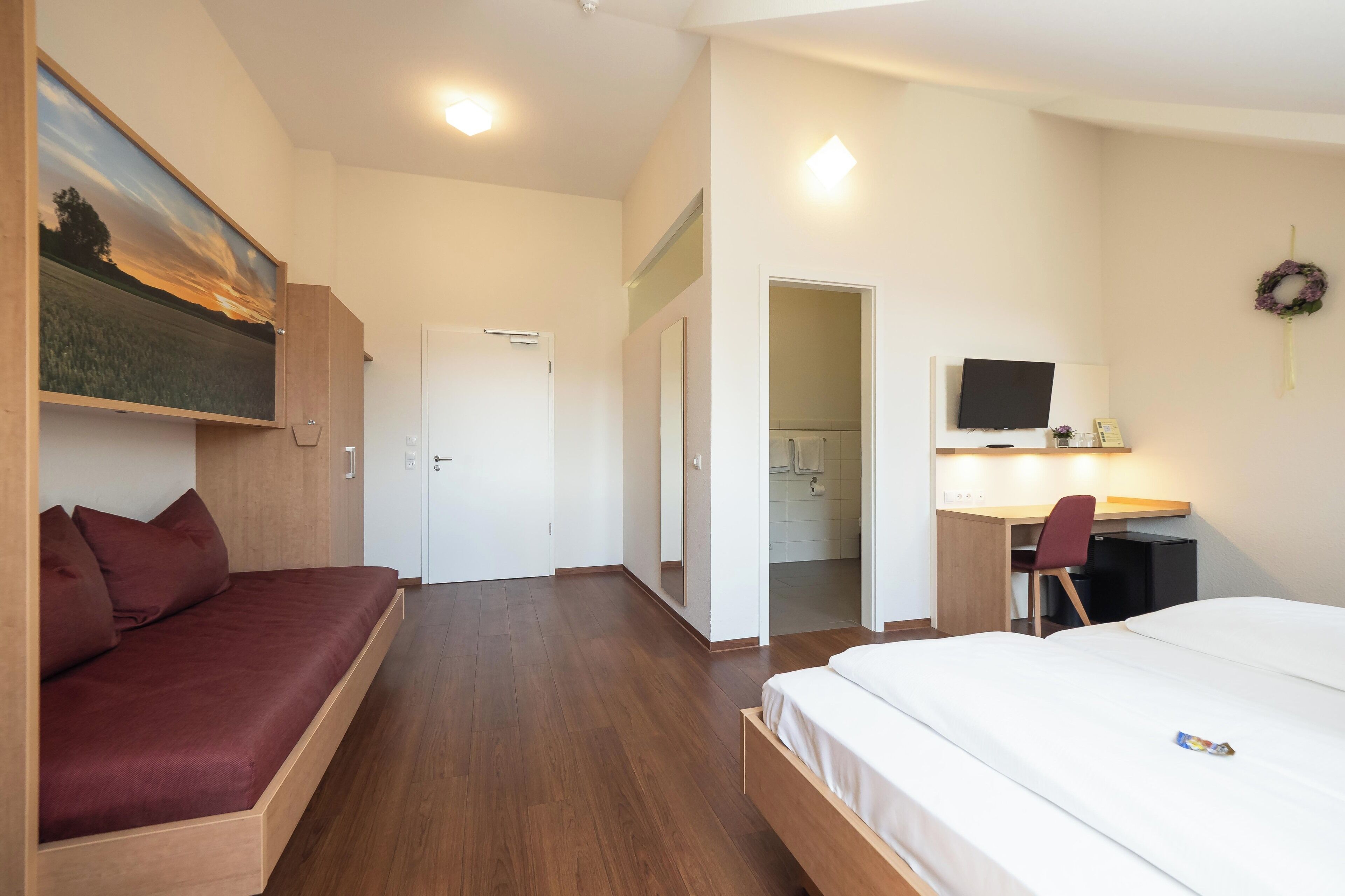 A modern hotel room with a bed, a sofa, a desk with a chair, a TV, and a painting on the wall.