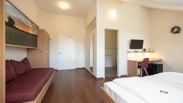 A modern hotel room with a bed, a sofa, a desk with a chair, a TV, and a painting on the wall.