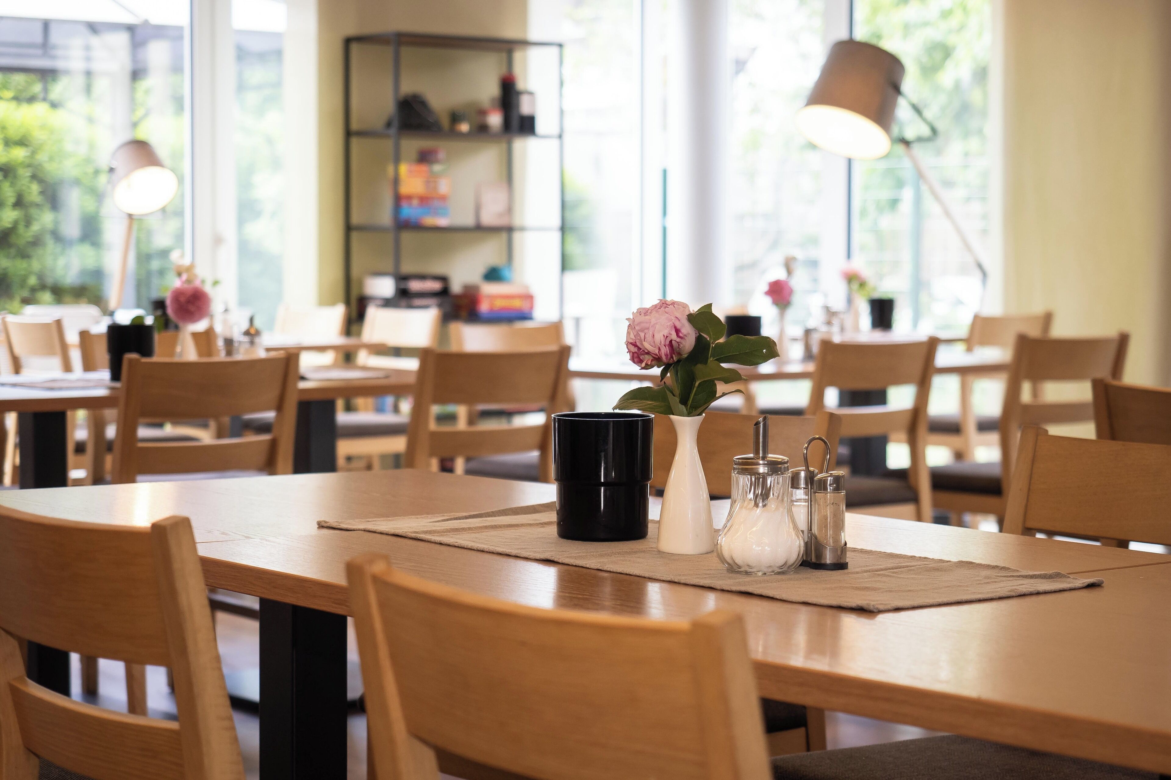 A dining area with wooden tables and chairs, a vase with a flower, salt and pepper shakers, and a black mug.