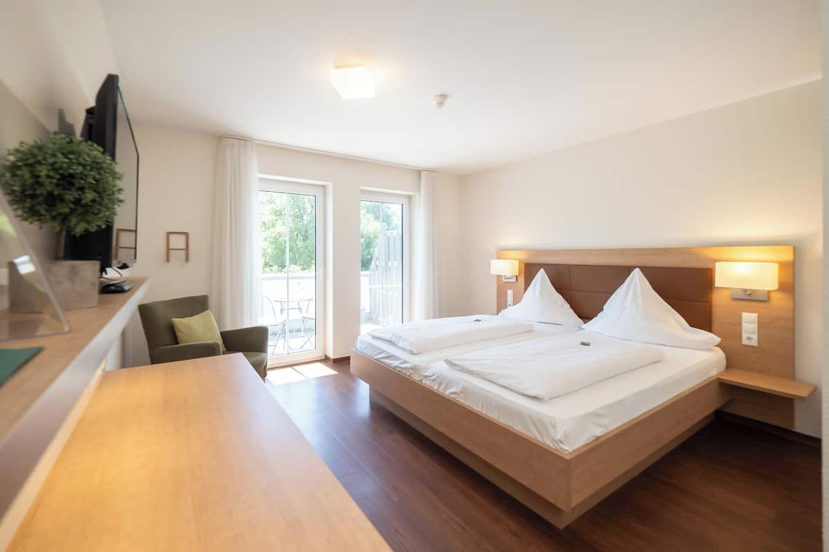 Een hotelkamer met een groot bed, een bureau en een stoel. Op het bureau staat een plant en er is een raam met gordijnen.