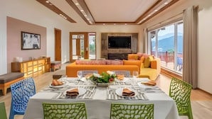 A modern living room with a sofa, dining table, and TV. There is a balcony with a view of the mountains. - amã Stays & Trails Kedar Villa, Mukteshwar (Mukteshwar)