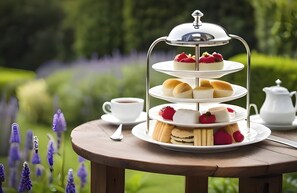 Un présentoir à trois niveaux, garni de diverses pâtisseries et de fruits, une théière blanche et une tasse de thé, le tout posé sur une table en bois dans un jardin.