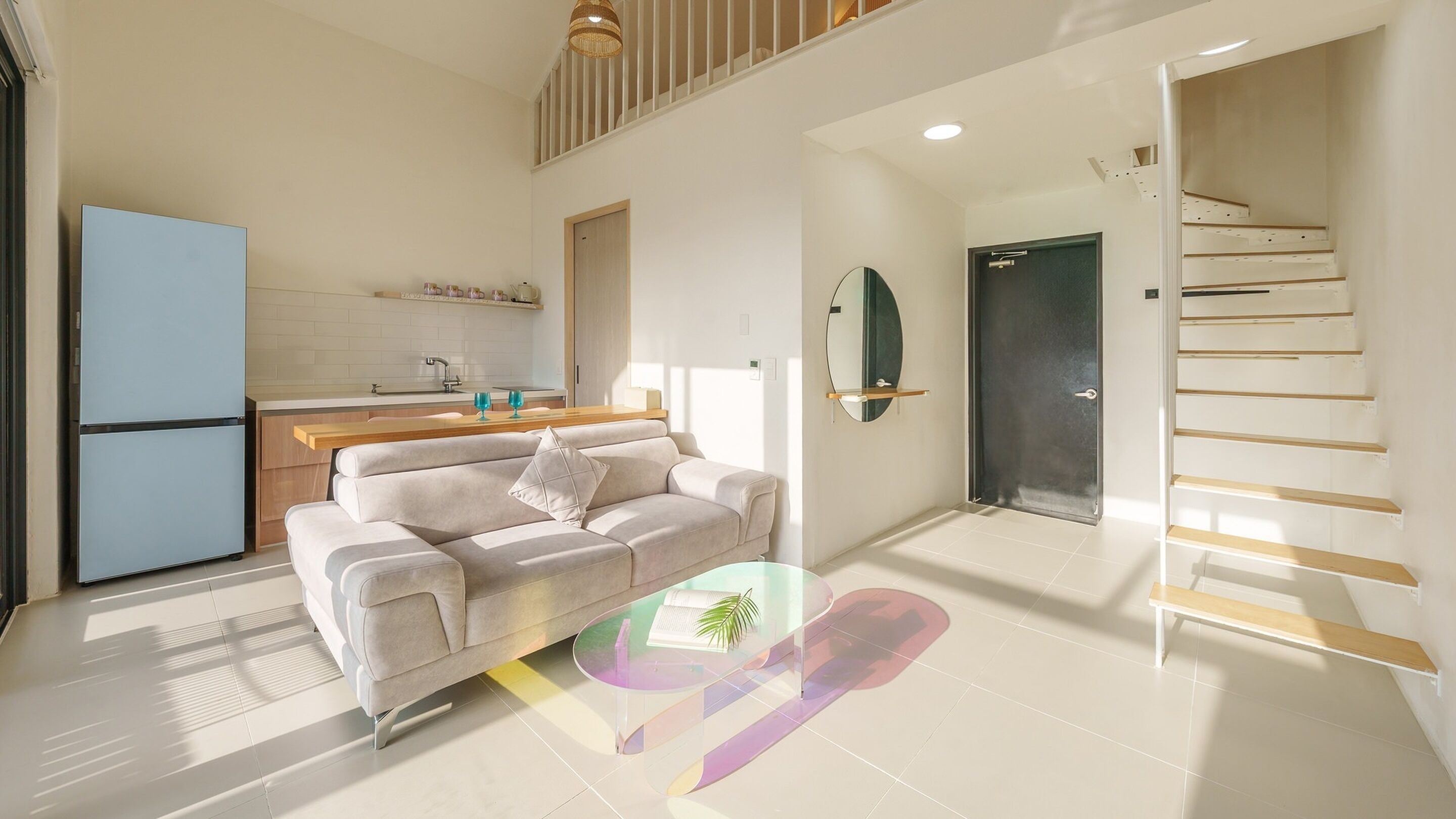 A modern living room with a sofa, a glass coffee table, and a kitchen area with a refrigerator and sink.