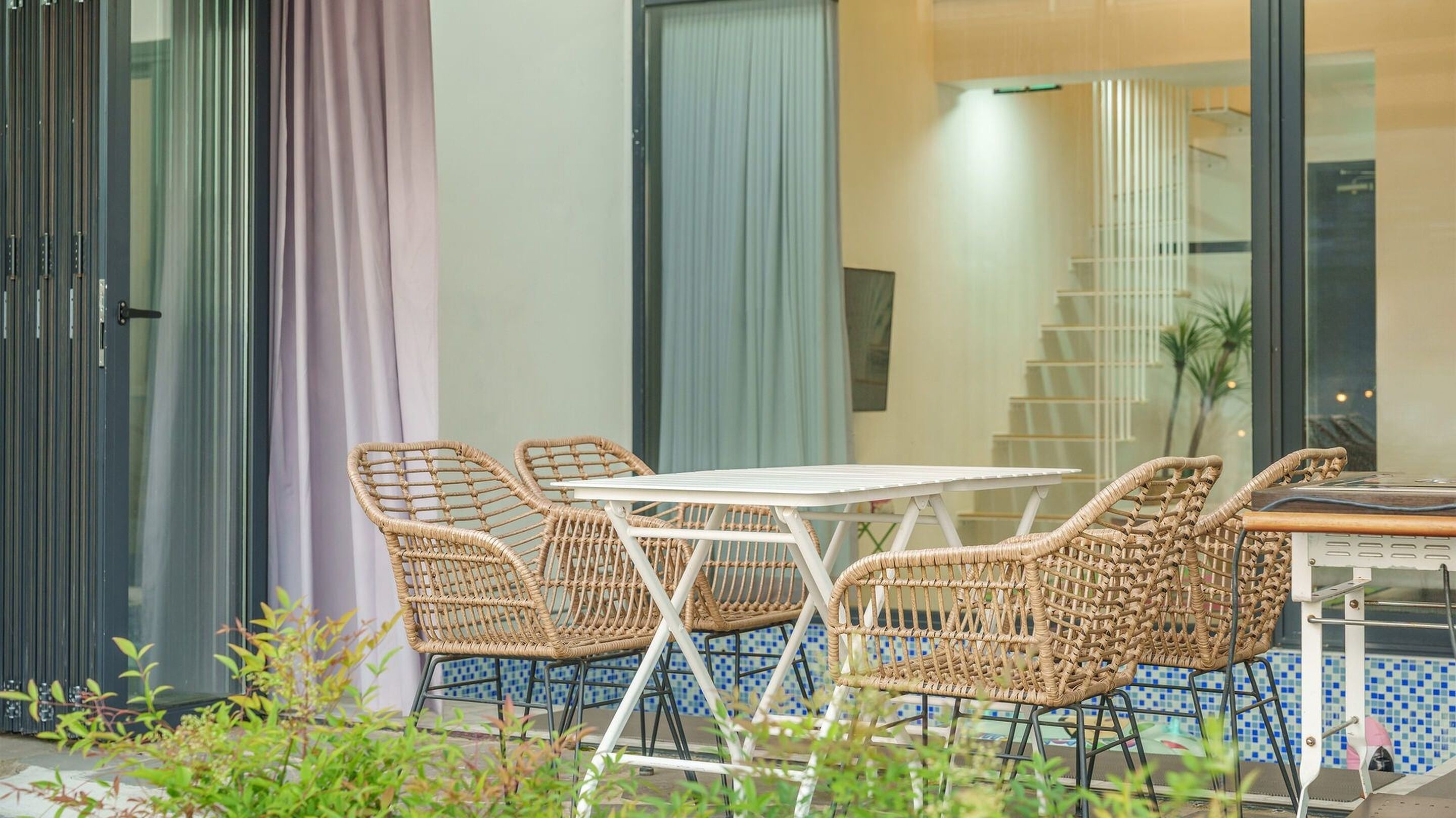 A modern outdoor seating area with wicker chairs and a white table, set against a building with large windows and a tiled floor.