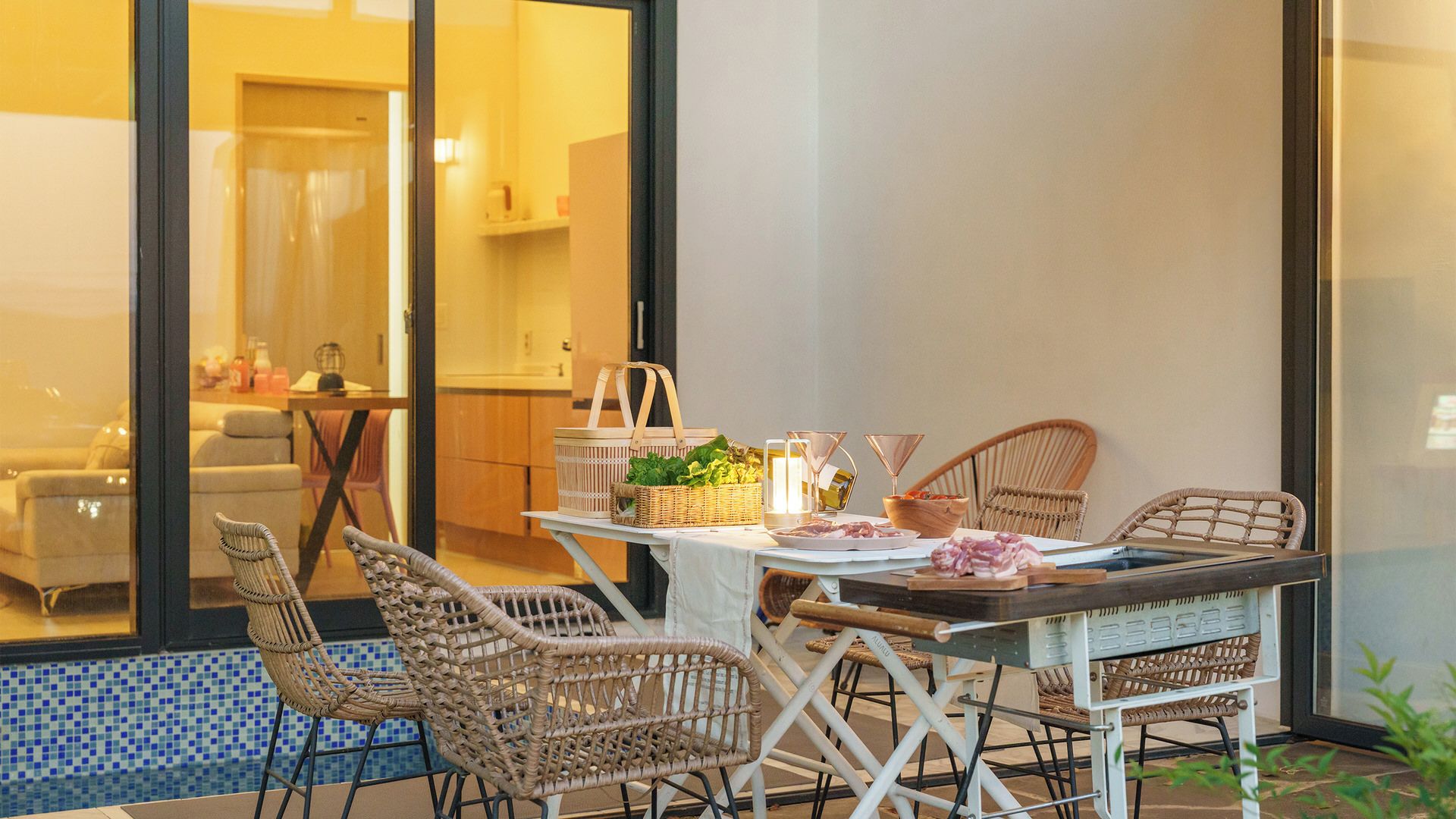 Un coin repas avec des chaises en osier, une table recouverte d’une nappe blanche et une vue sur une piscine, visible à travers de grandes portes vitrées.