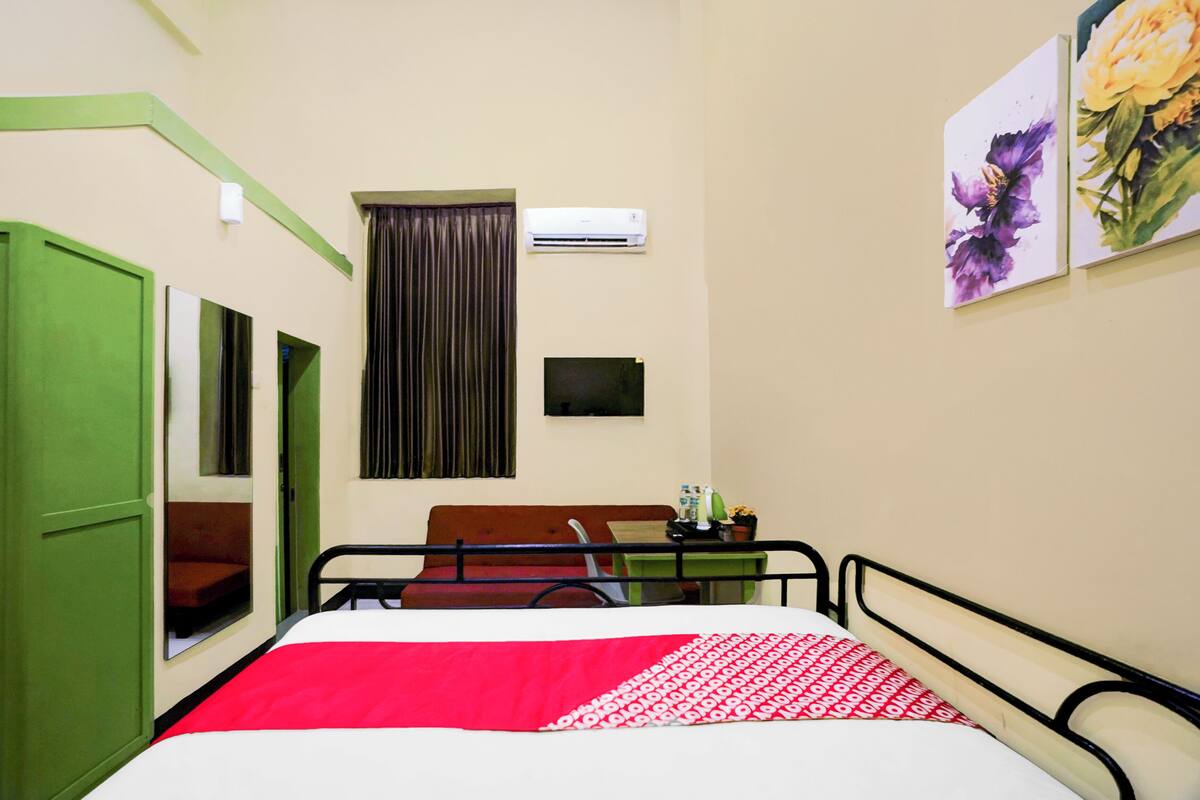A hotel room with a bed, a desk, a chair, a window with curtains, an air conditioner, and two framed pictures on the wall.