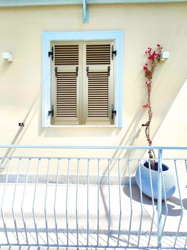 A balcony with a white railing, a window with wooden shutters, and a potted plant with pink flowers.