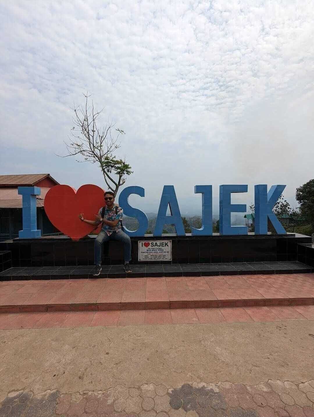 A large blue sign with "I ♥ SAJEK" and a red heart symbol, with a person sitting on a bench in front of it.