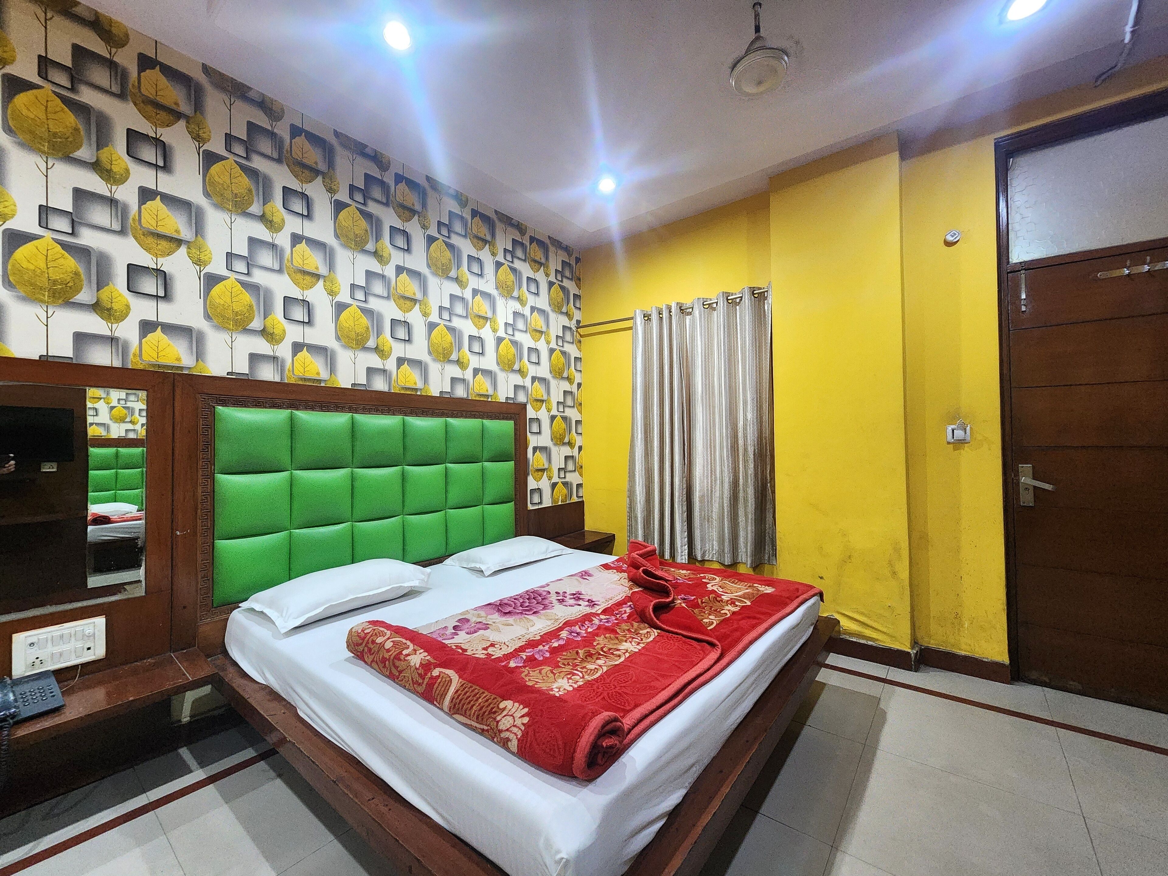 A hotel room with a green upholstered headboard, a bed with white linens and a red floral blanket, a patterned wall, and a yellow wall with a door.