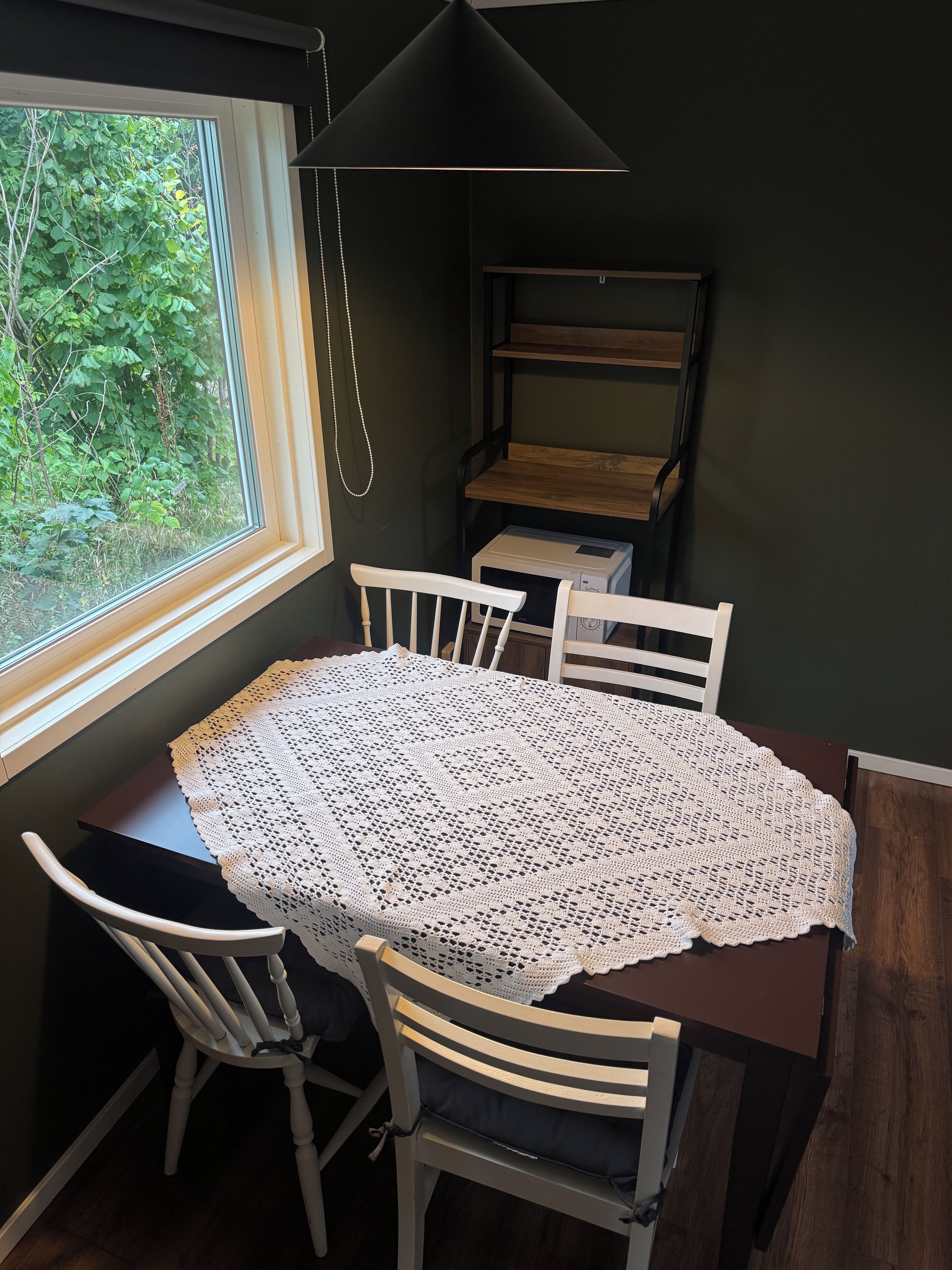Uma área de refeições com uma mesa coberta por um mantel branco de crochê, duas cadeiras brancas, uma luminária pendente preta e vista para o verde através de uma janela.