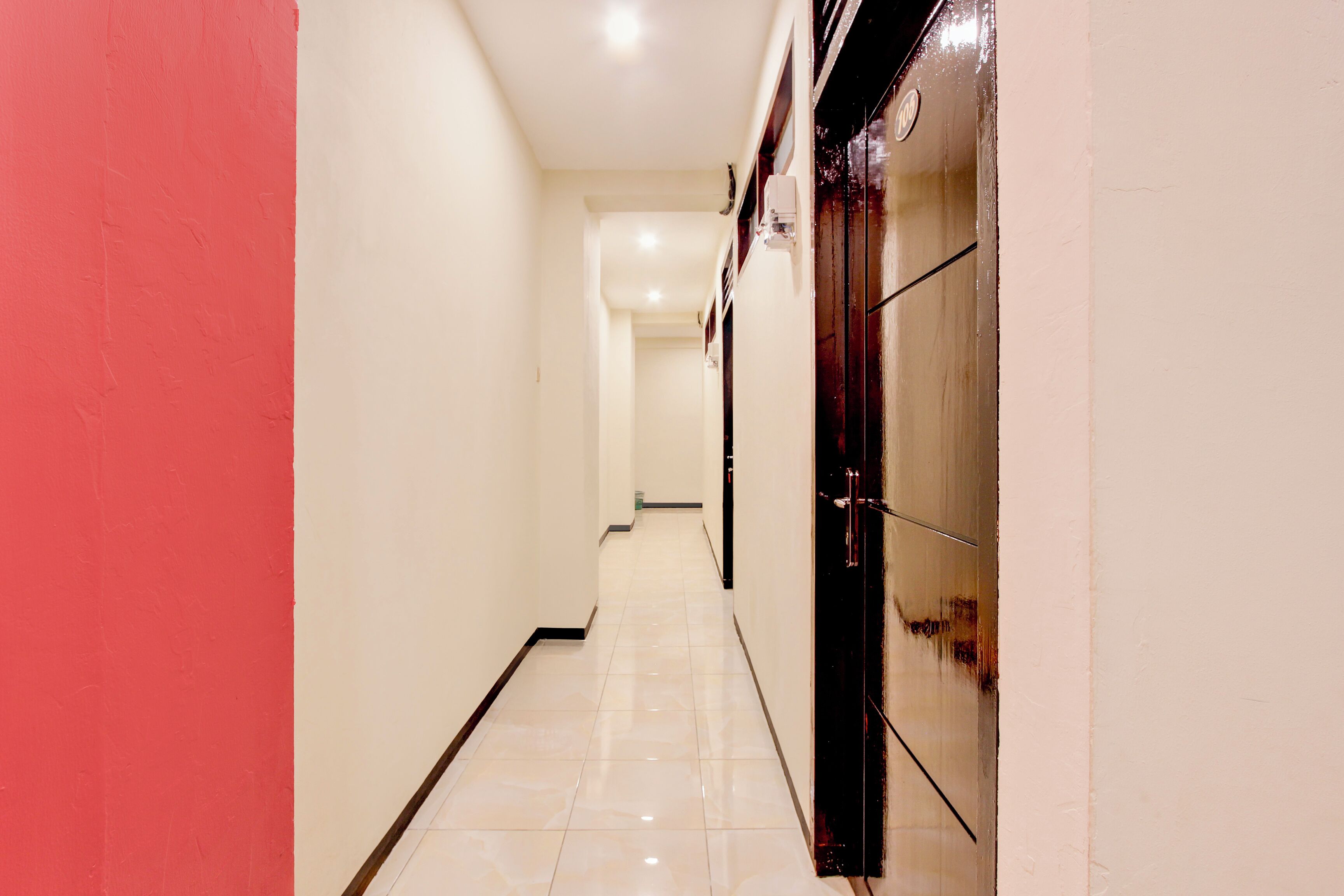 A corridor with a red wall on one side and a door with a glass panel on the other. The floor is tiled, and the walls are painted in neutral colors.