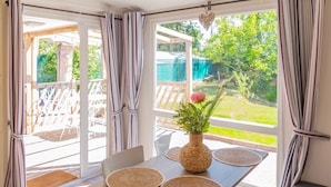 Een kamer met een gedekte tafel voor twee personen, een rieten vaas met groen en een uitzicht op een tuin door grote ramen.