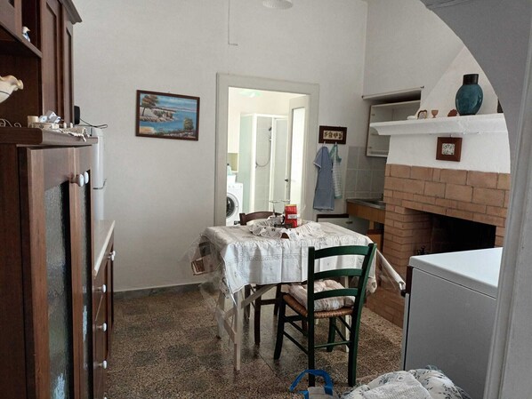 Een praktische keuken met een eettafel, stoelen, een koelkast, een schouw van bakstenen en een wasmachine op de achtergrond.