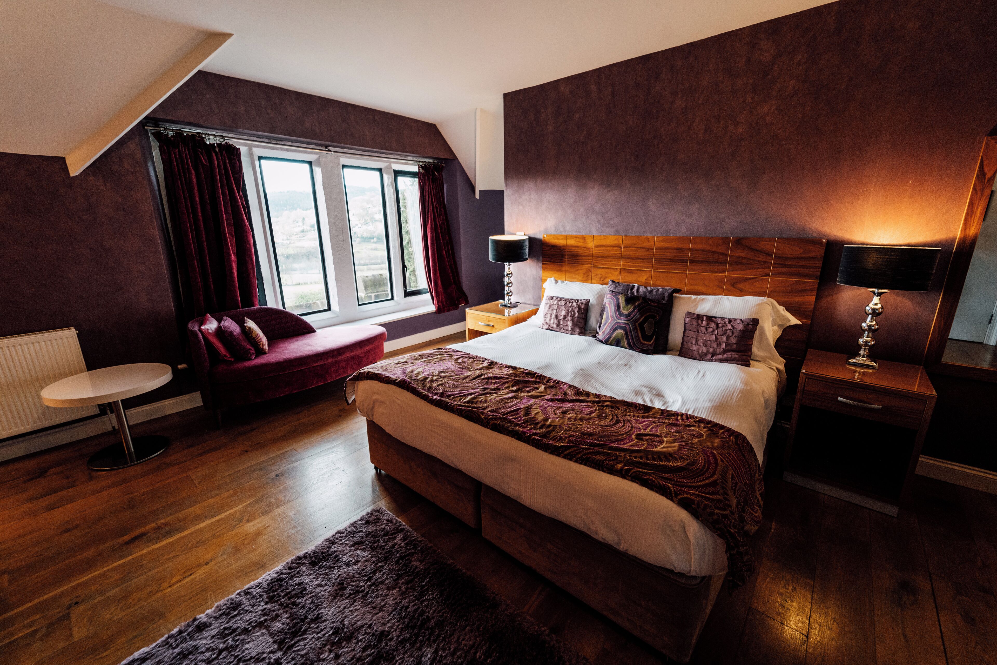 A modern bedroom with a large bed, a small round table, a red chair, and a view of the outdoors through a window.