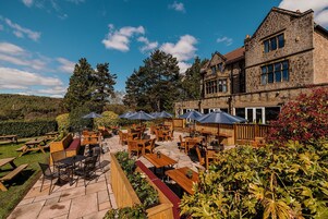 An outdoor patio with wooden tables and chairs, stone flooring, and a stone building in the background.