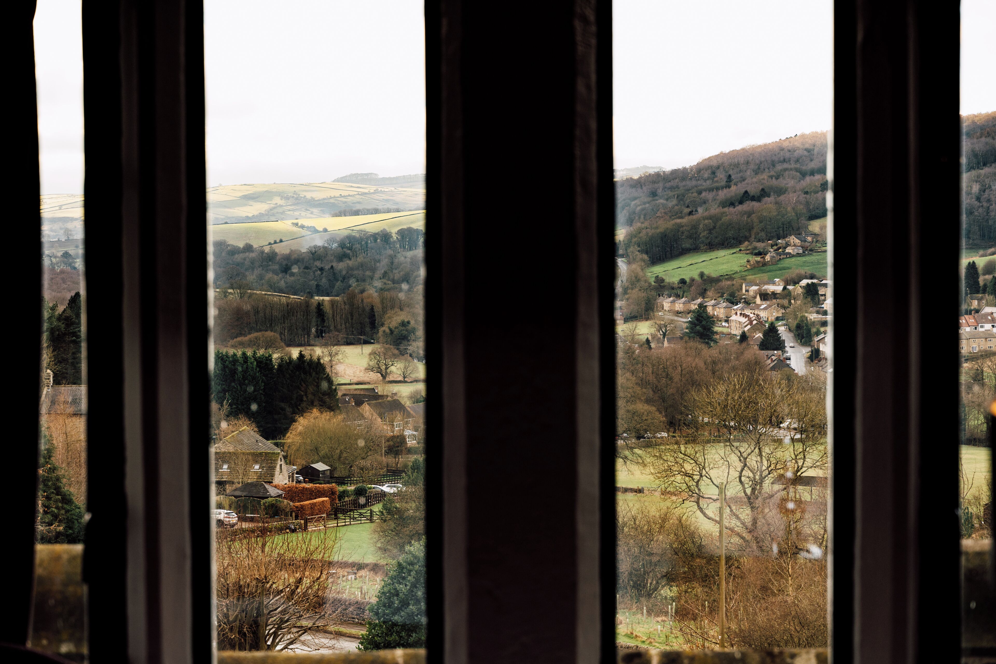 Vista a través de una ventana que muestra un paisaje rural con colinas ondulantes, un pequeño pueblo y una iglesia.
