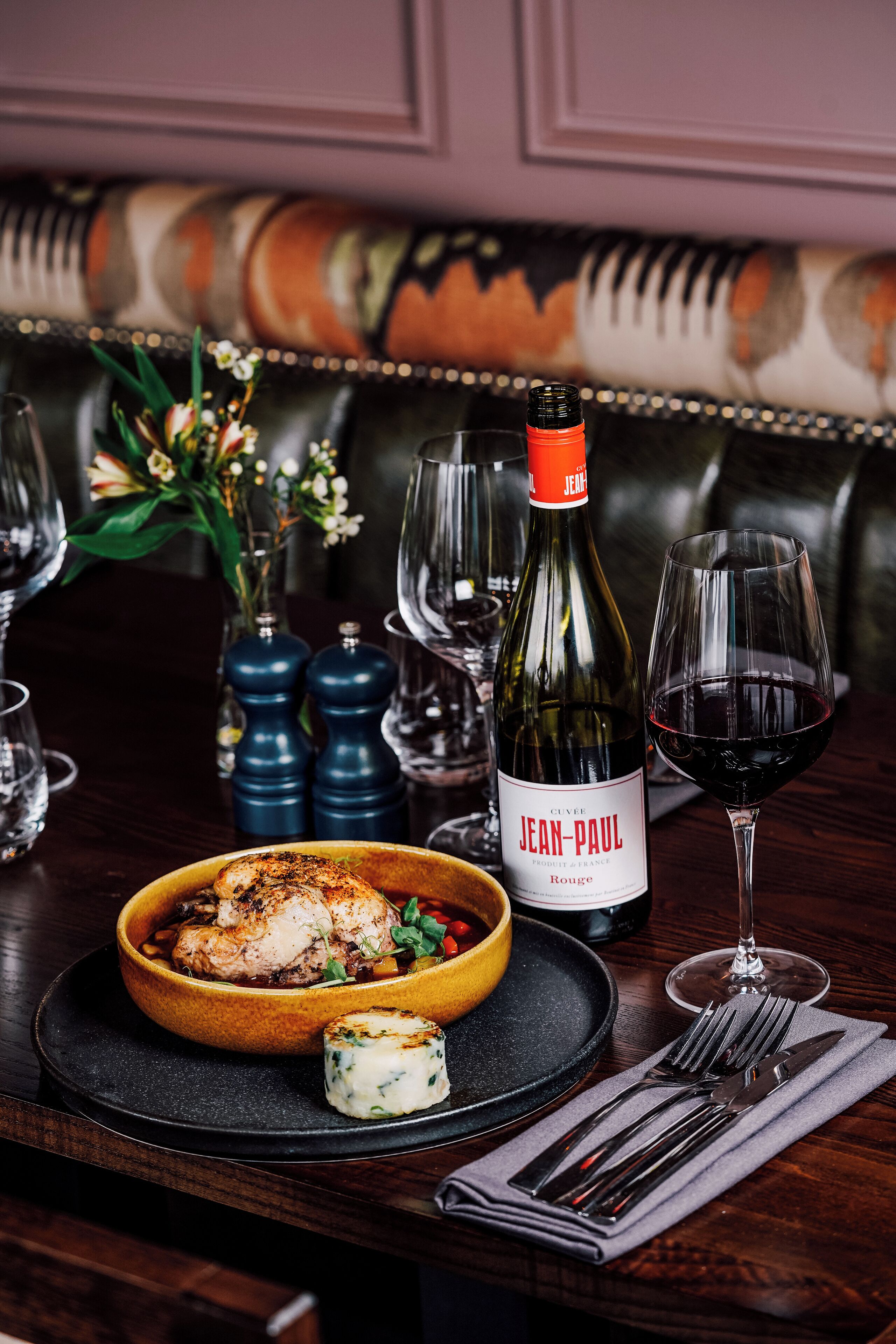 Una mesa de comedor con una botella de Jean-Paul, una copa de vino tinto, un plato de pollo y un pequeño recipiente con queso.