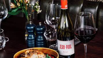 Una mesa de comedor con una botella de Jean-Paul, una copa de vino tinto, un plato de pollo y un pequeño recipiente con queso.