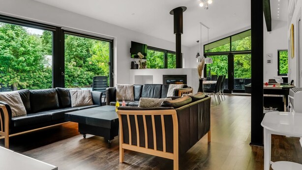 Una sala moderna con un sofá de cuero negro, una mesa de centro de madera y un comedor con una mesa y sillas blancas. Amplias ventanas ofrecen vistas a zonas verdes.