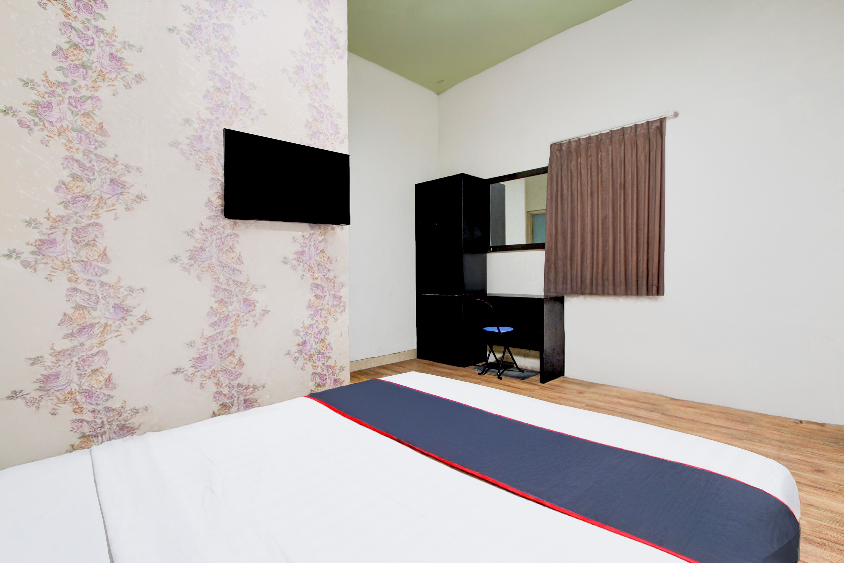 A bedroom with a bed, a television mounted on the wall, a desk with a chair, and a window with curtains.