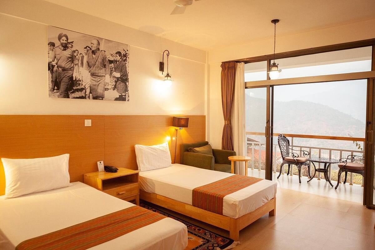 A hotel room with two beds, a balcony with chairs, and a large window offering a mountain view.