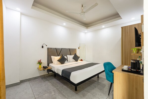 A modern hotel room with a bed, bedside table, chair, desk, and a ceiling fan. - Hotel Admire Inn Sec - 104 By ASPL (Noida)