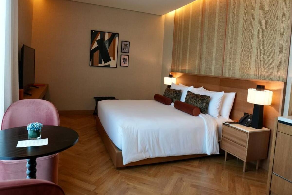 A hotel room with a large bed, a bedside table with a lamp, a small table with a vase, and a chair.