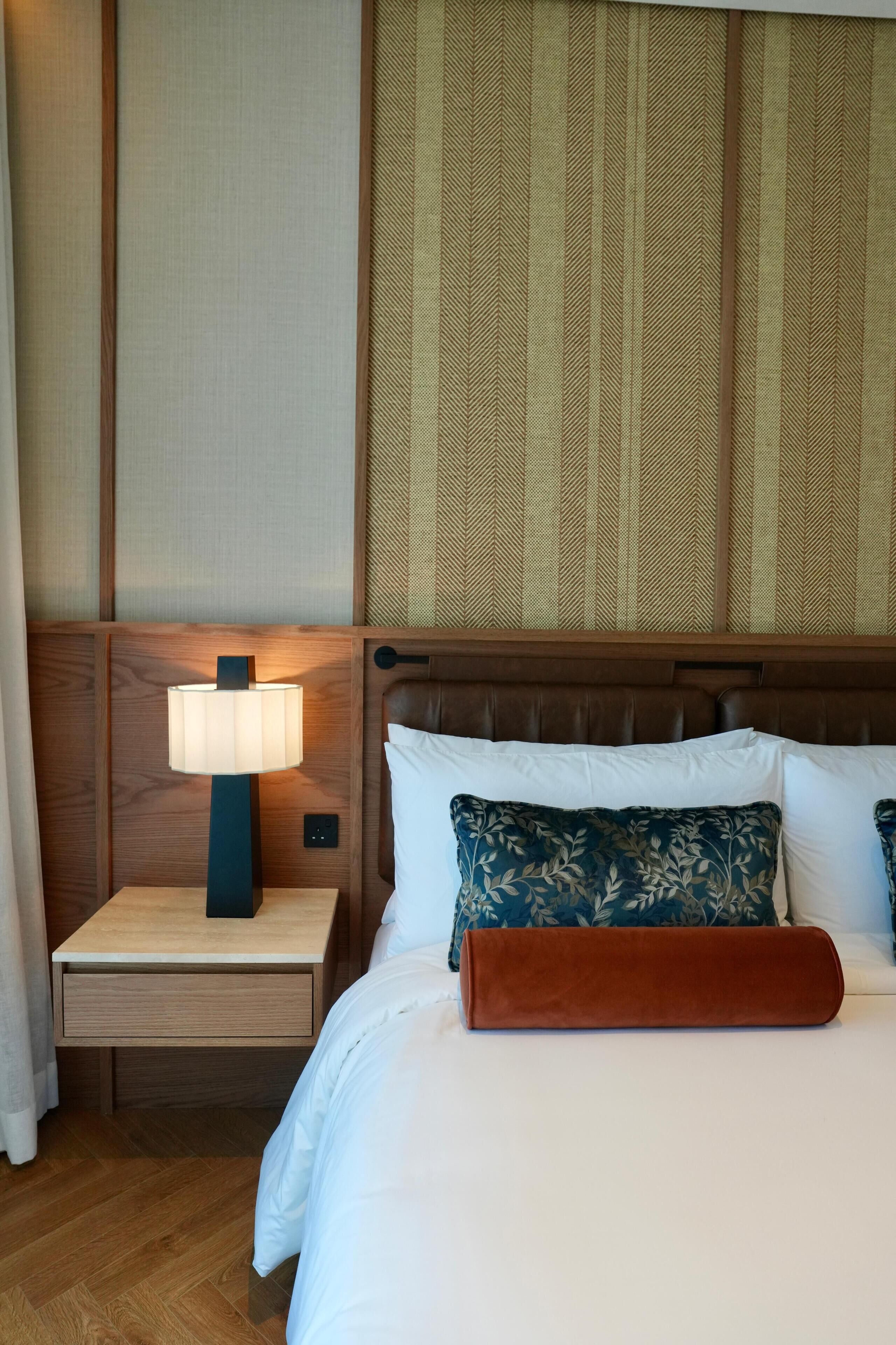 A hotel room with a wooden headboard, a nightstand with a lamp, a bed with white and blue pillows, and a brown throw blanket.