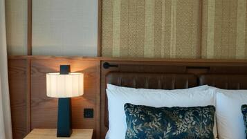 A hotel room with a wooden headboard, a nightstand with a lamp, a bed with white and blue pillows, and a brown throw blanket.
