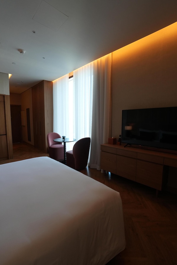 A hotel room with a large bed, a TV on a wooden stand, a small table with a chair, and a window with curtains.