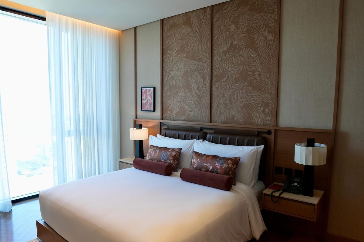 A modern hotel room with a large bed, wooden headboard, and a view of the outdoors through a large window.